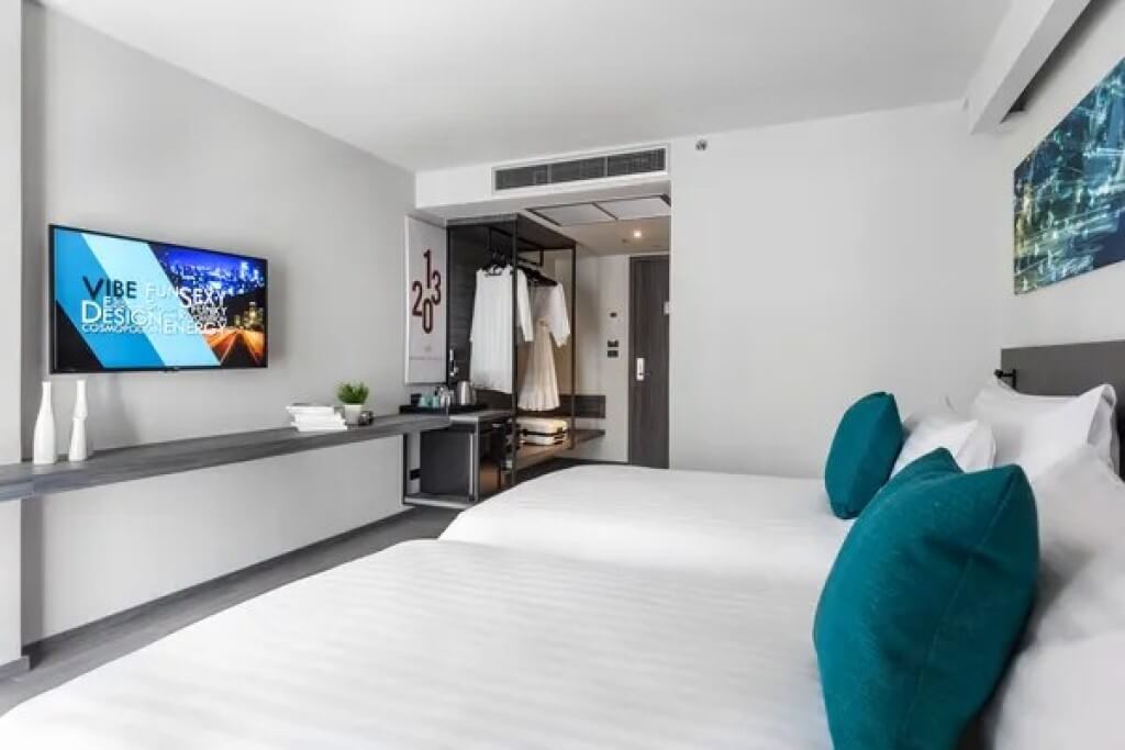 Cross Vibe Bangkok Sukhumvit twin room seen from window side facing beds, wall-mounted wardrobe rack, and in-room amenity shelf area